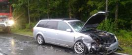 Srebrny samochód osobowy stoi na drodze i ma rozbity przód, za nim stoi samochód gaśniczy.