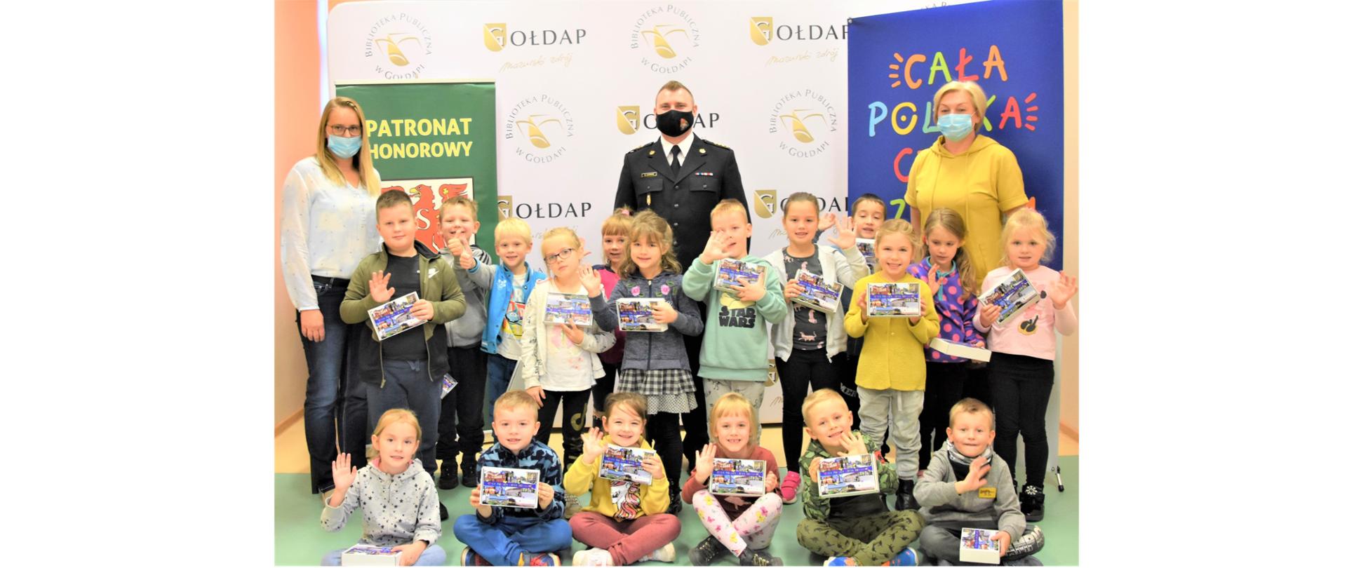 Zdjęcie przedstawia osiemnaścioro dzieci klasy 0 „C” ze Szkoły Podstawowej nr 3 im. Tadeusza Kościuszki w Gołdapi, które były słuchaczami bajek oraz od lewej: Panią Dyrektor Biblioteki Publicznej w Gołdapi, st. kpt. Michała Herman i Panią wychowawcę klasy, na tle m.in. baneru akcji „Cała Polska czyta dzieciom”
