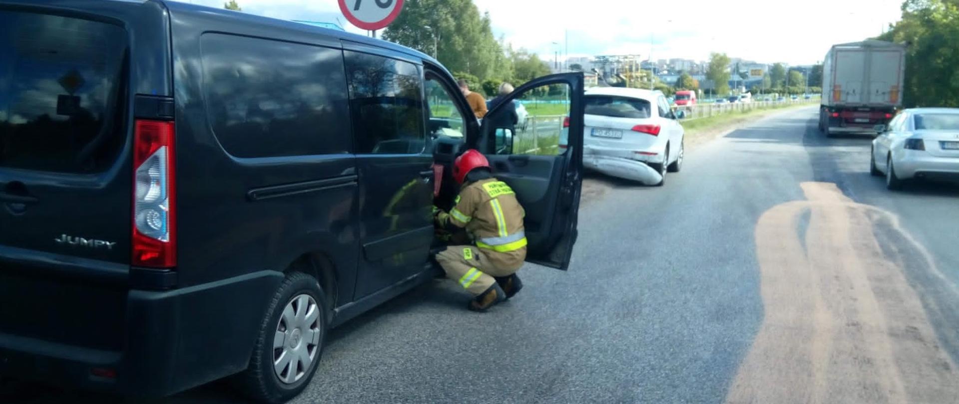 Zdjęcie przedstawia perspektywę ulicy Łódzkiej w Kielcach. Na pierwszym planie, zepchnięty na pobocze stoi pierwszy z uszkodzonych aut. W jego otwartych drzwiach znajduje się strażak. Przed nim stoi również na poboczu drugi z biorących udział w kolizji pojazdów. Prawą stroną obywa się ruch drogowy choć na środku widać znaczną plamę płynów eksploatacyjnych posypanych Damolinem.
