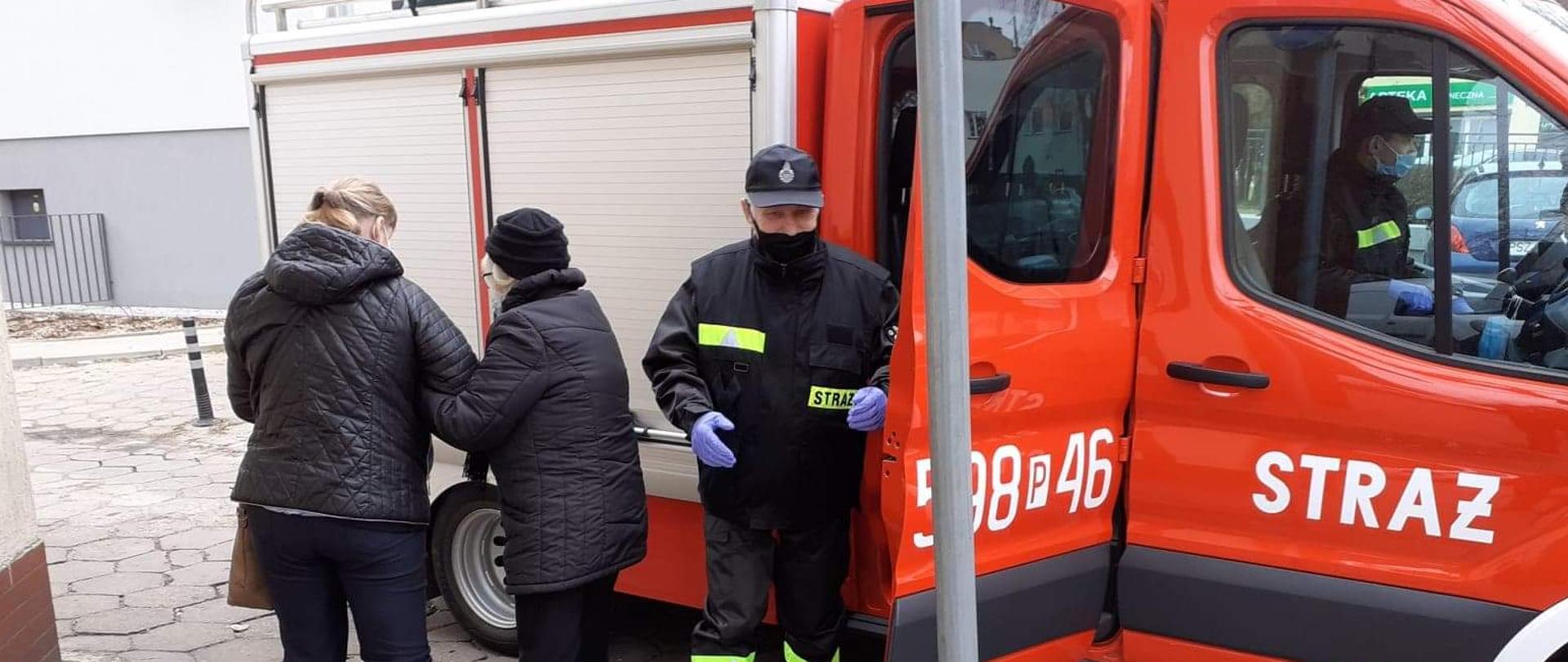 Widać samochód straży pożarnej, strażaka oraz dwie osoby