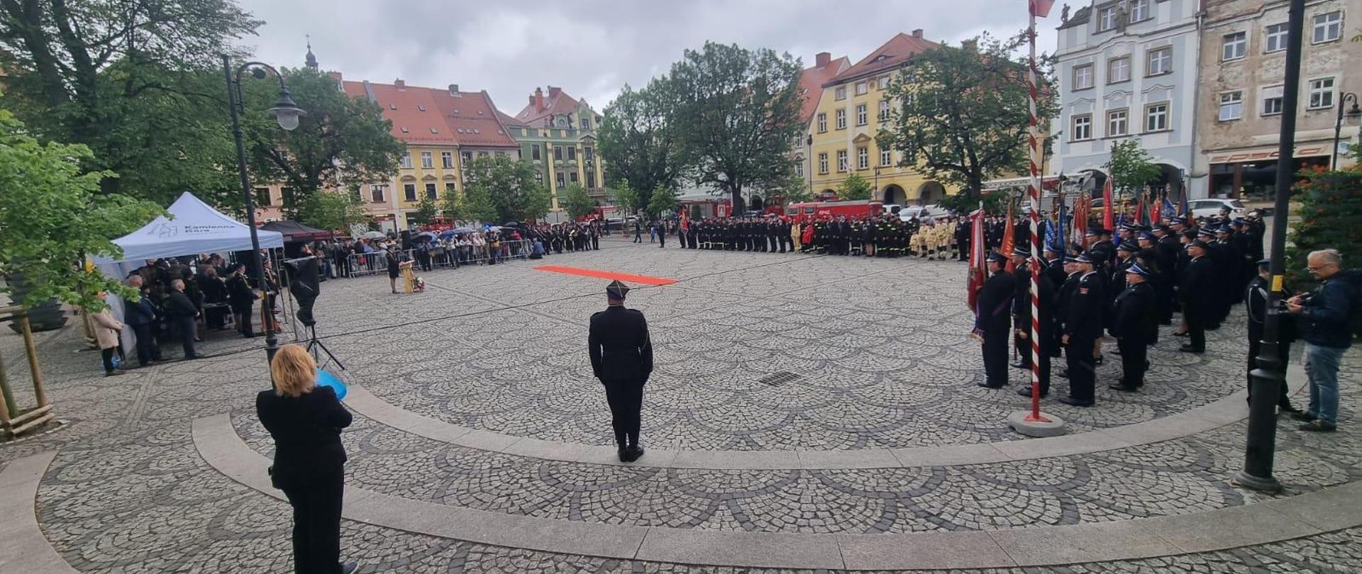 Obchody Dnia Strażaka na rynku miasta Kamienna Góra. Strażacy ustawieni na placu. Po lewej stronie namiot w którym stoją zaproszeni goście.