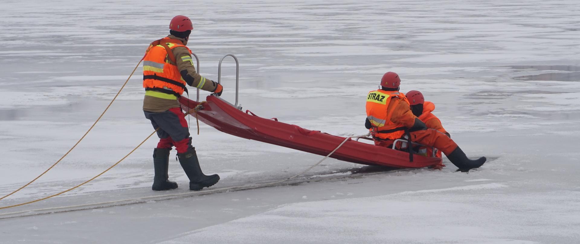 Dwóch strazaków ewakuuje osobę poszkodowana z przerębla prz użyciu sań lodowych obaj sa asekurowani prz uzyciu linek ratowniczych