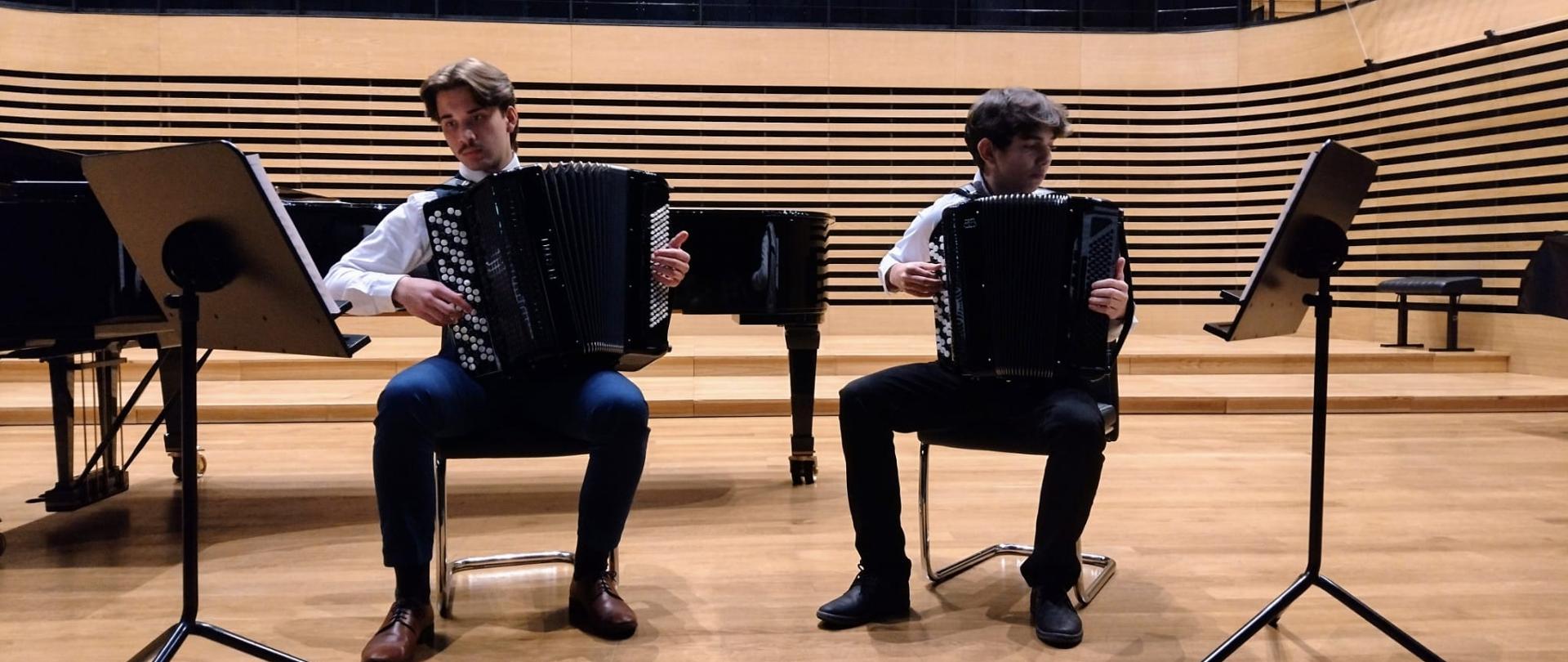 Dóch nastolatków sidzie obok siebie na krzesłach i grają na akordeonach, po bokach są ustawione pulpity w które spoglądają.