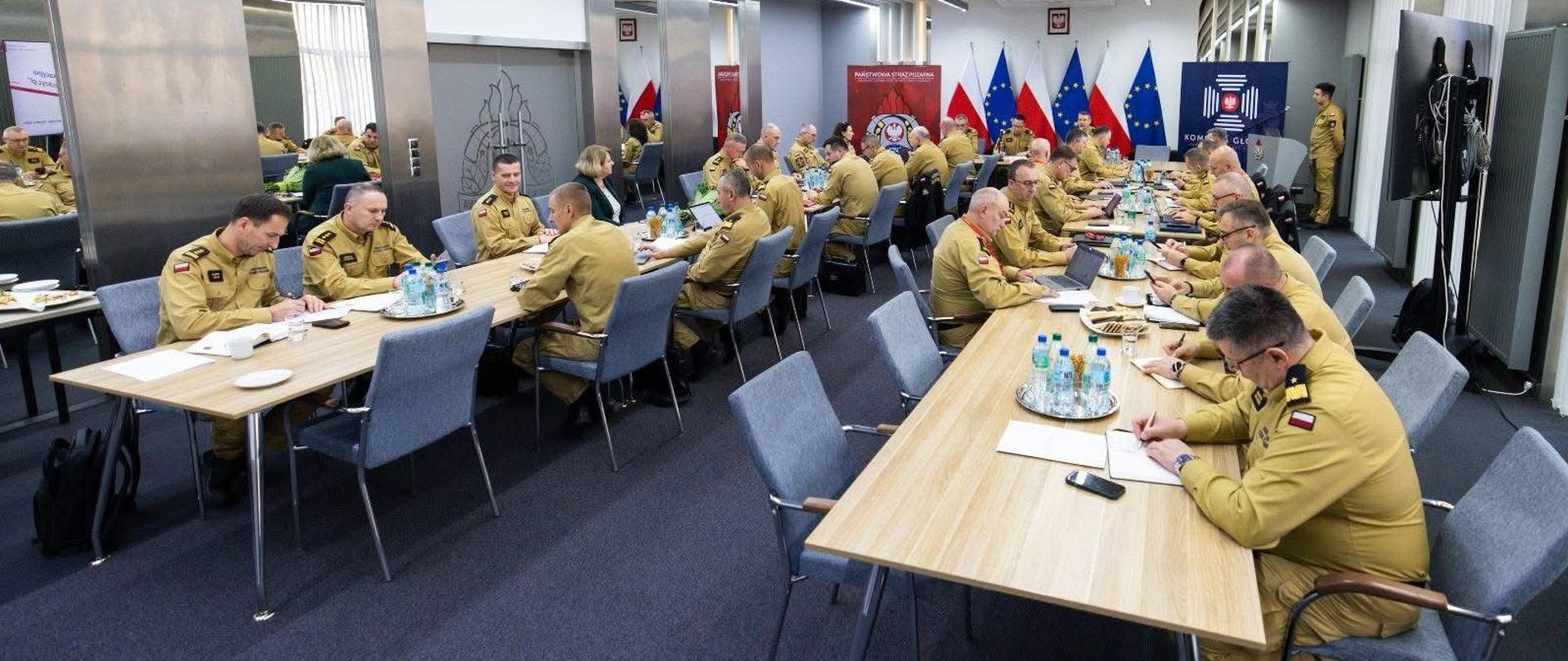 Na zdjęciu widać dużą, nowoczesną salę konferencyjną. Przy kilku długich, prostokątnych stołach siedzi kilkadziesiąt osób ubranych w jednakowe, piaskowe mundury służbowe. Siedzą na krzesłach, pochylają się nad dokumentami, notują lub pracują na laptopach. Na stołach znajdują się butelki z wodą, szklanki, notesy i telefony. W tle sali stoją flagi Polski oraz Unii Europejskiej, a także banery instytucjonalne. Pomieszczenie jest dobrze oświetlone, z jasnym sufitem i nowoczesnym wyposażeniem technicznym. 