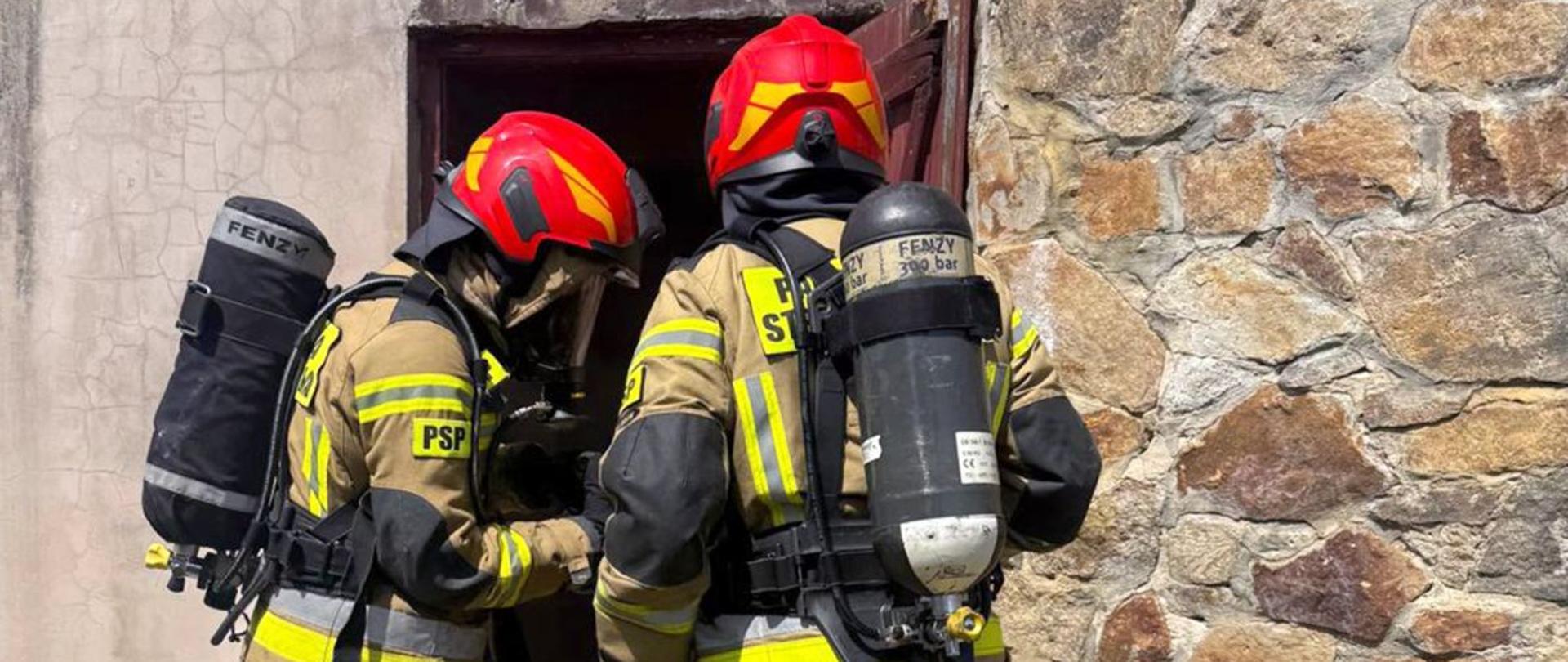 Zdjęcie przedstawia dwóch strażaków w ubraniach specjalnych z założonymi aparatami powietrznymi i butlami na plecach. Strażacy stoją przed wejściem do piwnicy.