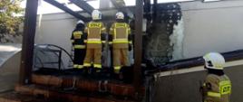 Strażacy w ubraniach bojowych prowadzą działania gaśnicze przy altanie, której konstrukcja i ściana budynku zostały nadpalone.