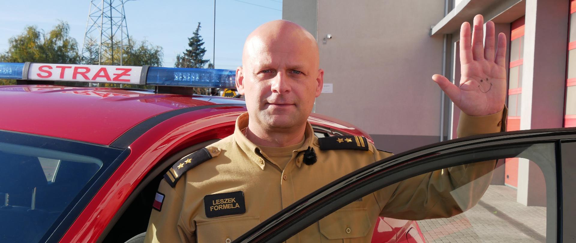 Komendant Powiatowy Państwowej Straży Pożarnej w Lęborku stoi przy otwartych drzwiach samochodu. Trzyma uniesioną do góry lewą dłoń, na której ma namalowaną uśmiechniętą buźkę, symbol solidarności z popularyzacją Światowego Dnia Adopcji. 