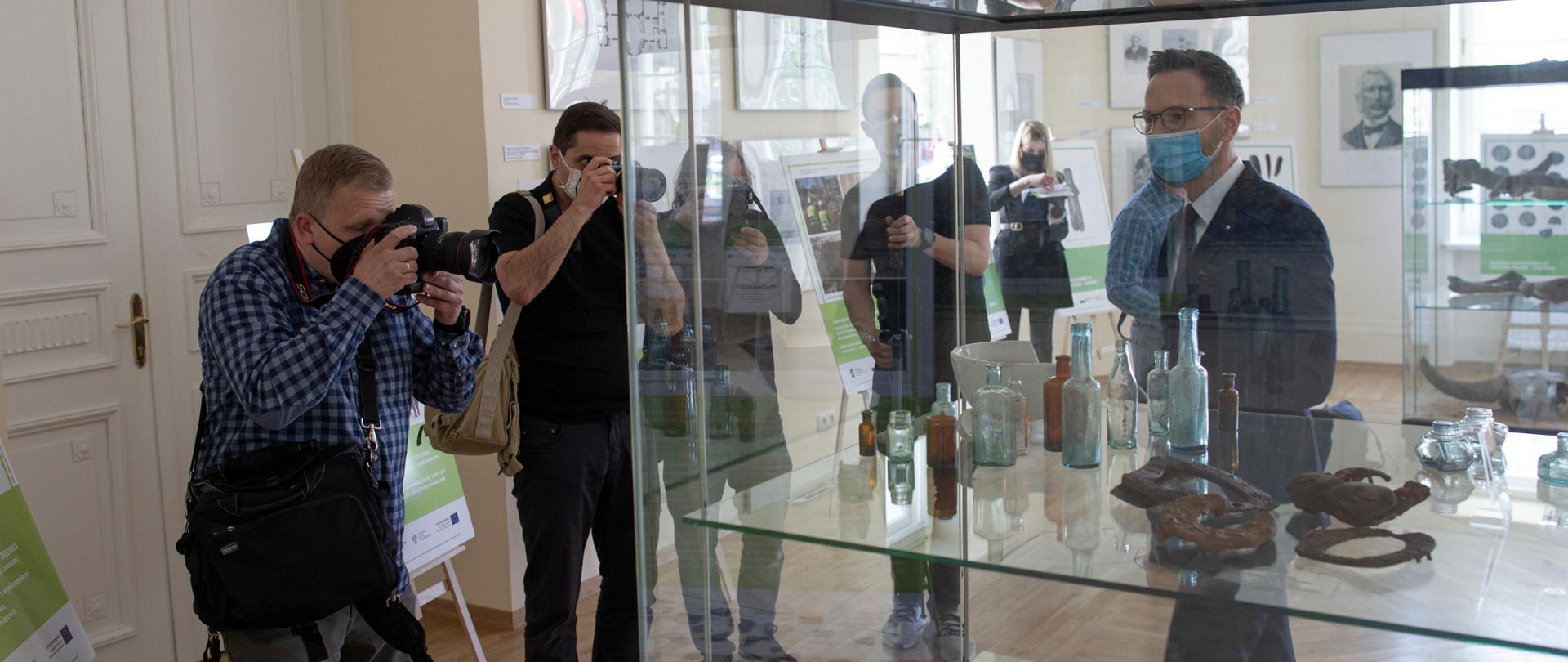 przed wyeksponowanymi w szklanej gablocie znaleziskami stoi wiceminister Waldemar Buda, którego fotografuje dwóch reporterów