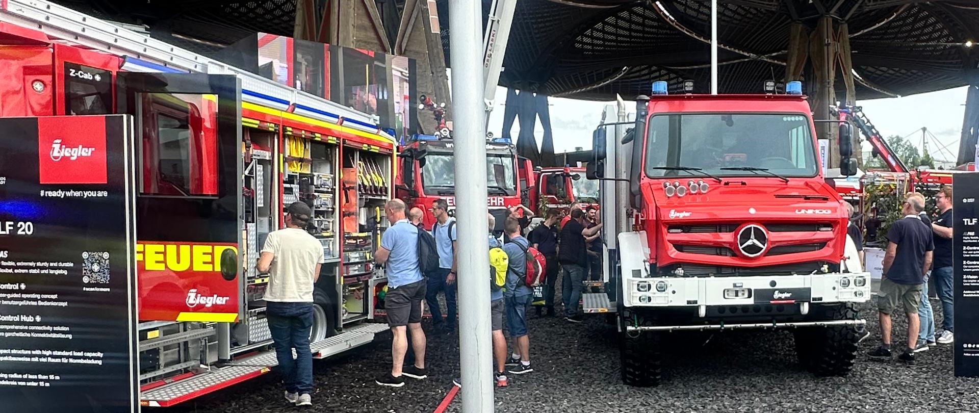 Zwiedzający oglądają specjalistyczne pojazdy pożarnicze prezentowane przez producentów podczas targów INTERSCHUTZ 2026. W centralnej części zdjęcia znajduje się czerwony samochód ratowniczy z podnośnikiem koszowym, ustawiony na stoisku wystawowym.