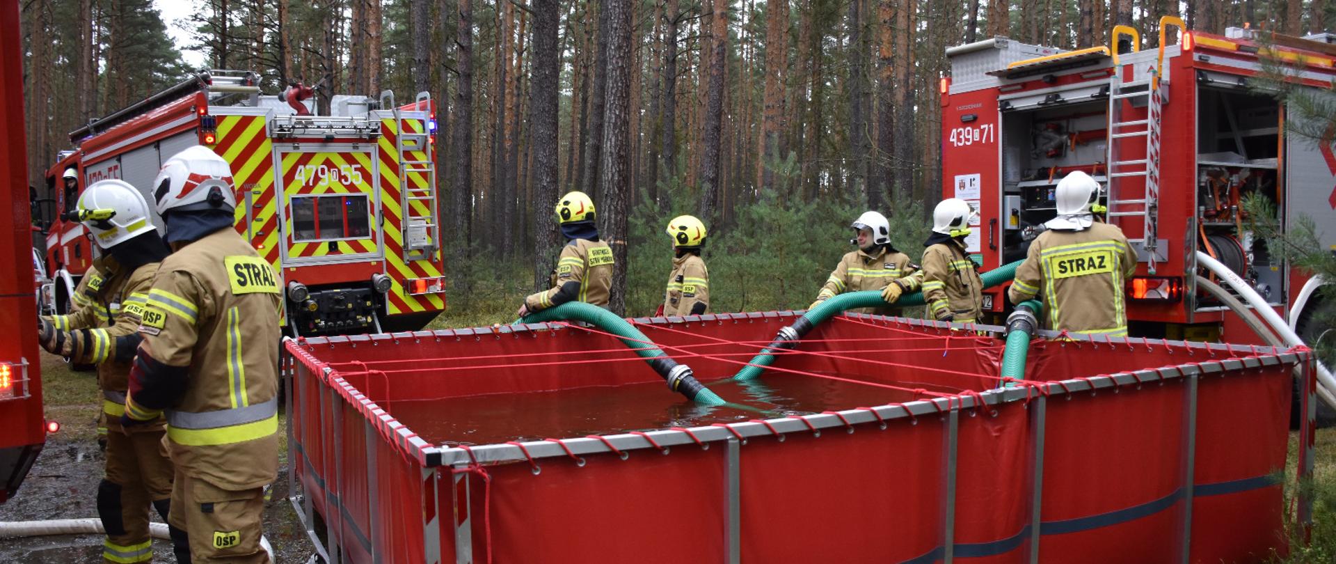 Strażacy napełniają przenośny zbiornik wodny w lesie przy użyciu węży i sprzętu gaśniczego podczas ćwiczeń „Las Pludry 2025”. W tle dwa wozy strażackie.