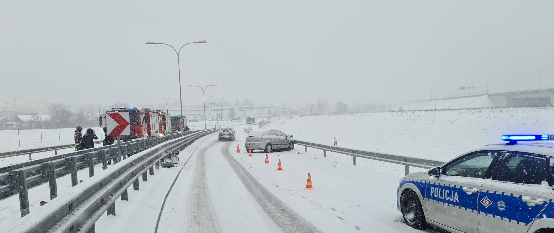 Na zdjęciu widoczny jest fragment trasy szybkiego ruchu S7. Na prawym pasie drogi na pierwszym planie znajduje się radiowóz policyjny. Przed nim znajduje się samochód osobowy w kolorze białym, który uderzył przodem w barierę energochłonną. Pojazd oznakowany jest pomarańczowymi pachołkami. Po lewej stronie na drugim pasie jezdni widać pojazdy pożarnicze