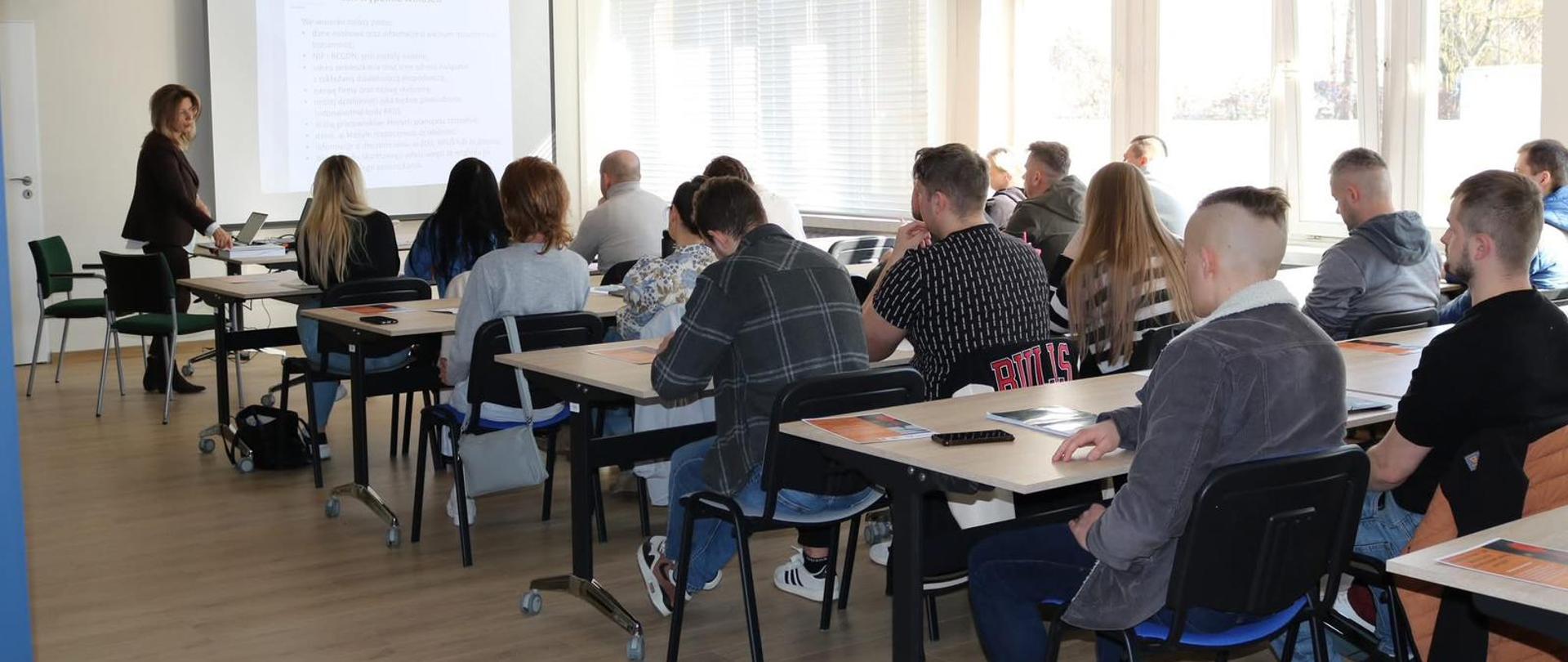 Grupa ludzi siedzi przy ławkach w sali szkoleniowej 