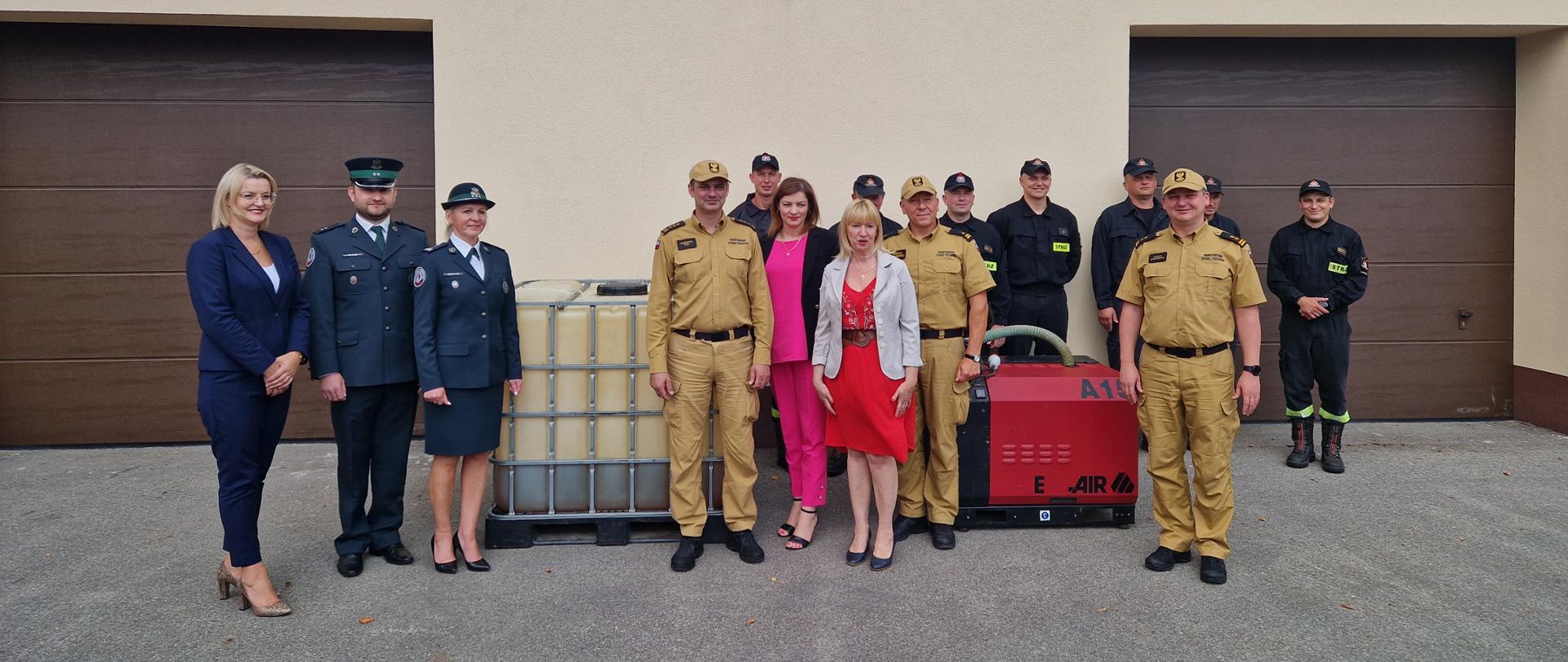 Zdjęcie przedstawia strażaków, funkcjonariuszy Krajowej Administracji Skarbowej oraz Urzędników przekazujących sprzęt i wyposażenie wołomińskim strażakom. Zdjęcie wykonane jest na tle garażu.