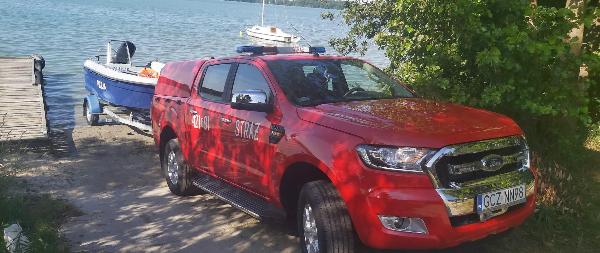 Na miejscu wodowania nad jeziorem Rychnowskim stoi strażacki samochód operacyjny Ford Ranger, który woduje policyjną łódź motorową.
