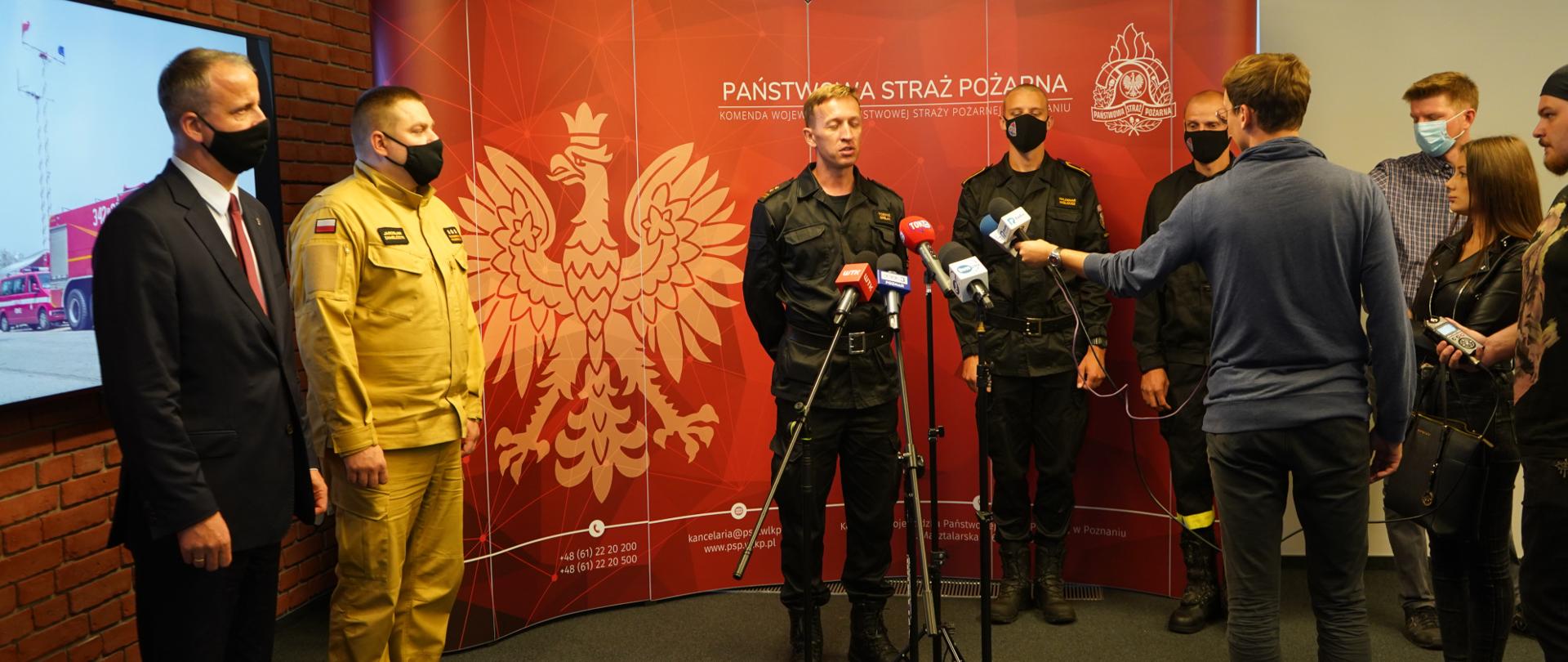 grupa strażaków stoi w szeregu, przed nimi mikrofony i redaktorzy, w tle czerwony baner i telewizor