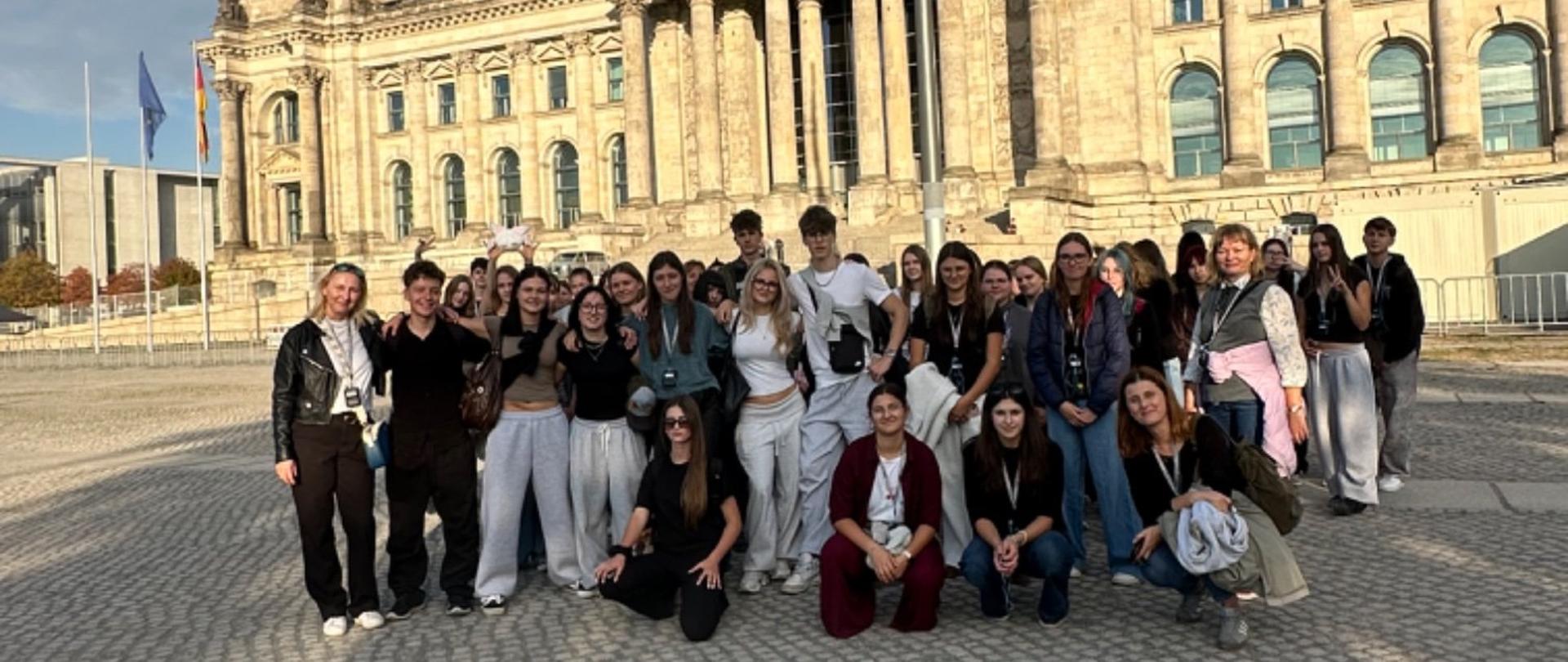 Grupa uczniów pozuje do wspólnego zdjęcia przed budynkiem Reichstagu w Berlinie. Nad placem widoczne są flagi Niemiec i zachmurzone niebo.