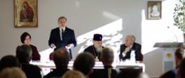 Wiceminister Edukacji Narodowej – Maciej Kopeć przemawia podczas konferencji. Obok siedzi arcybiskup Sawa – Prawosławny Metropolita Warszawski i całej Polski. W tle dwa obrazy na białej ścianie.