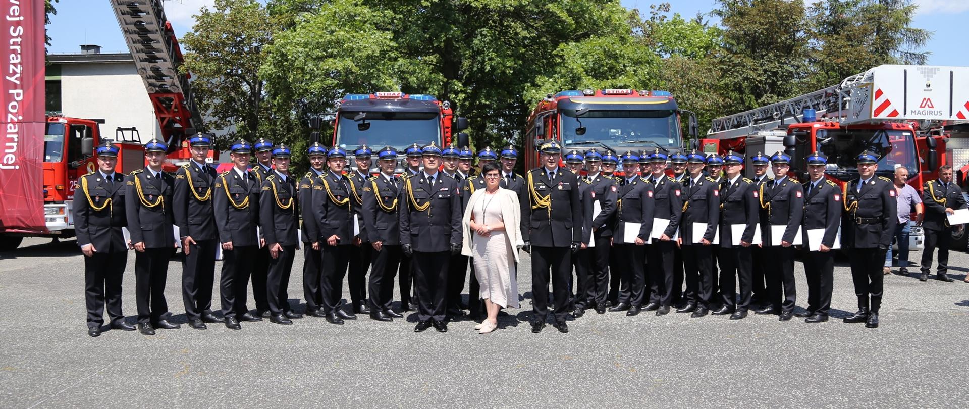 Grupowe zdjęcie absolwentów oraz uczestników promocji w towarzystwie Zastępcy Komendanta Głównego PSP, za nimi ustawione samochody pożarnicze