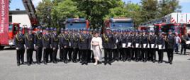Grupowe zdjęcie absolwentów oraz uczestników promocji w towarzystwie Zastępcy Komendanta Głównego PSP, za nimi ustawione samochody pożarnicze