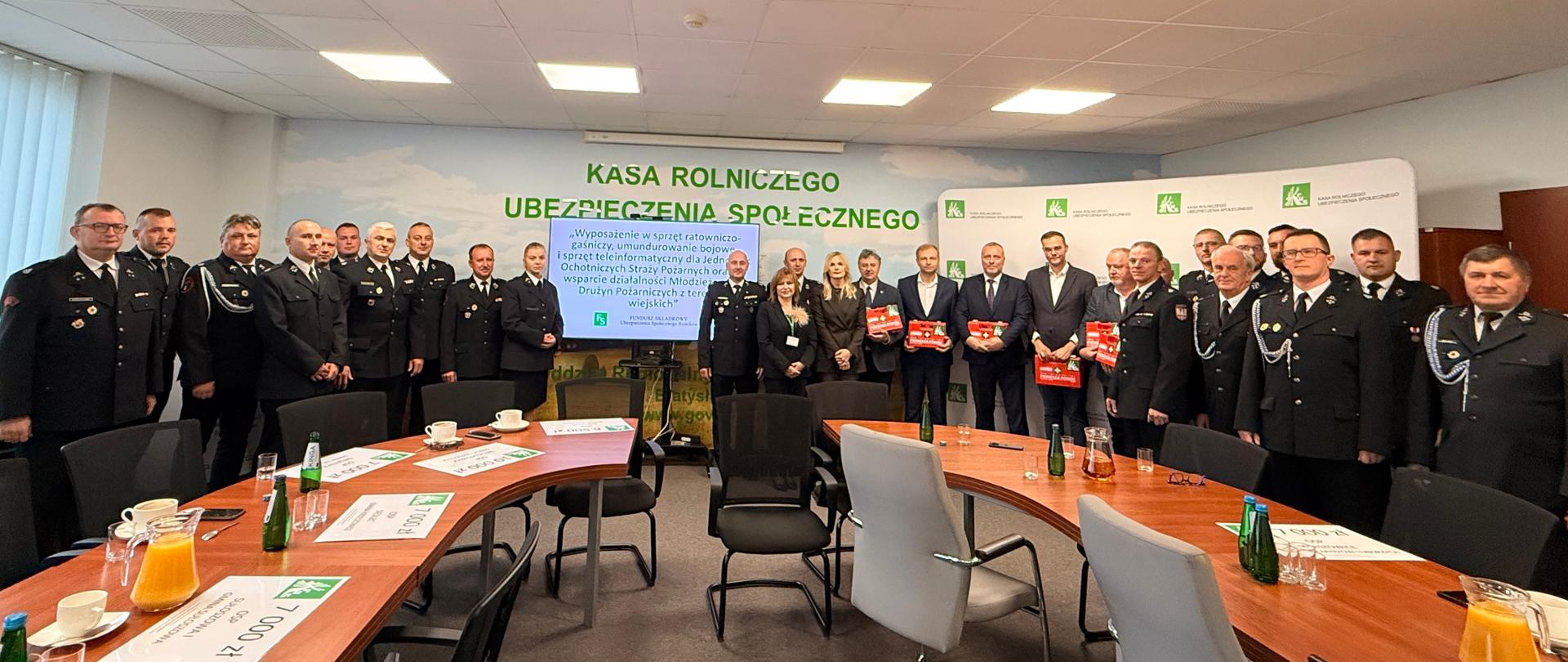 W sali konferencyjnej na tle ekranu i banerów z logo KRUS pozuje do zdjęcia ok. dwudziestu mężczyzn w mundurach OSP, W środku grupy stoją dwie kobiety i sześciu mężczyzn w garniturach, którzy trzymają w rękach czerwone apteczki.
