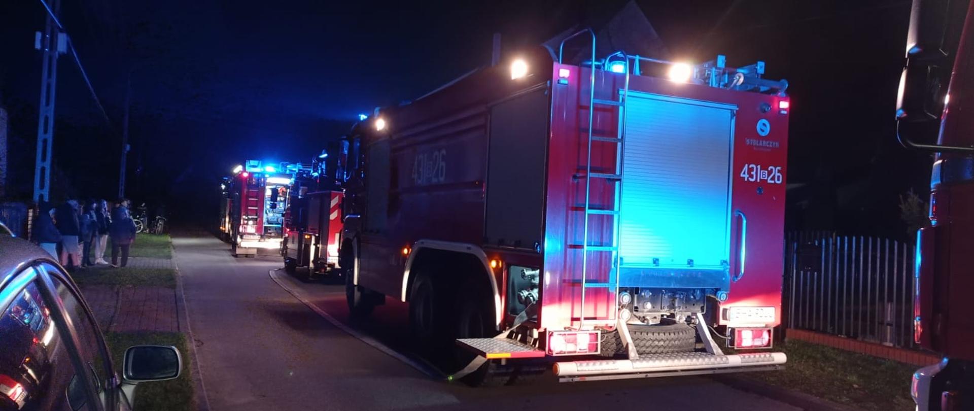 W nocy na drodze pomiędzy zabudowaniami stoją trzy samochody pożarnicze z włączonymi sygnałami świetlnymi.