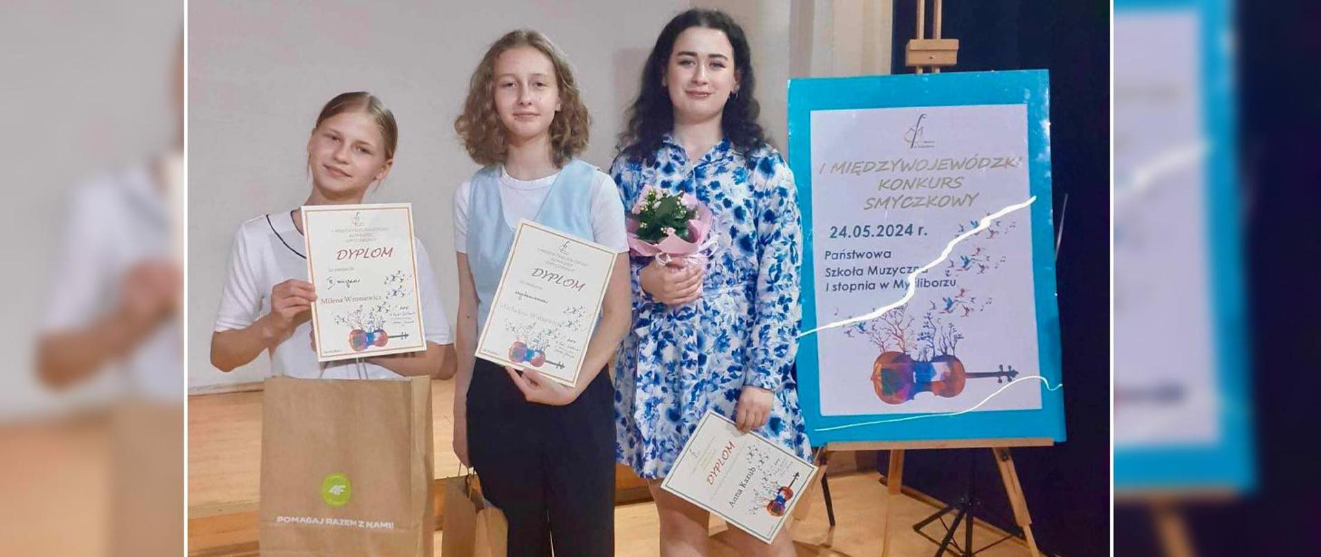 Na zdjęciu dwie uczennice i nauczycielka stoją obok banera I Międzywojewódzkiego Konkursu Smyczkowego. Wszystkie trzymają w rękach dyplomy. 