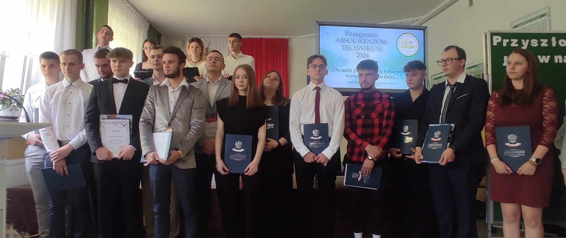 Portret grupowy absolwentów technikum rocznika 2024. Młodzi ludzie w eleganckich strojach prezentują otrzymane dyplomy, stojąc na tle szkolnej dekoracji.