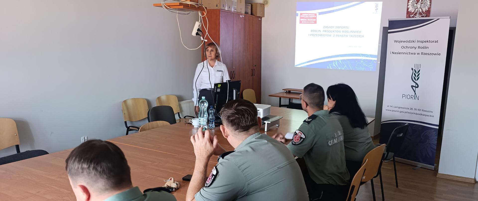 W sali przy dużym stole siedzą tyłem 4 osoby. Mają na sobie zielone koszule od munduru. W rogu stołu stoi kobieta, ma przed sobą komputer. Na ścianie wyświetla się prezentacja. Po prawej stronie stoi baner instytucji. 