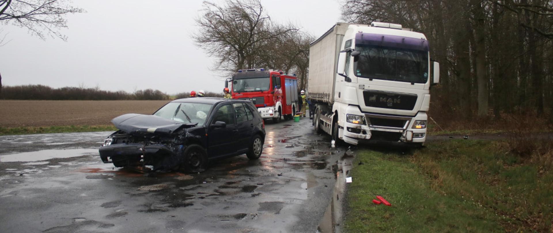 Zdjęcie przedstawia uszkodzony samochód osobowy i ciężarowy. W tle stoi samochód pożarniczy.