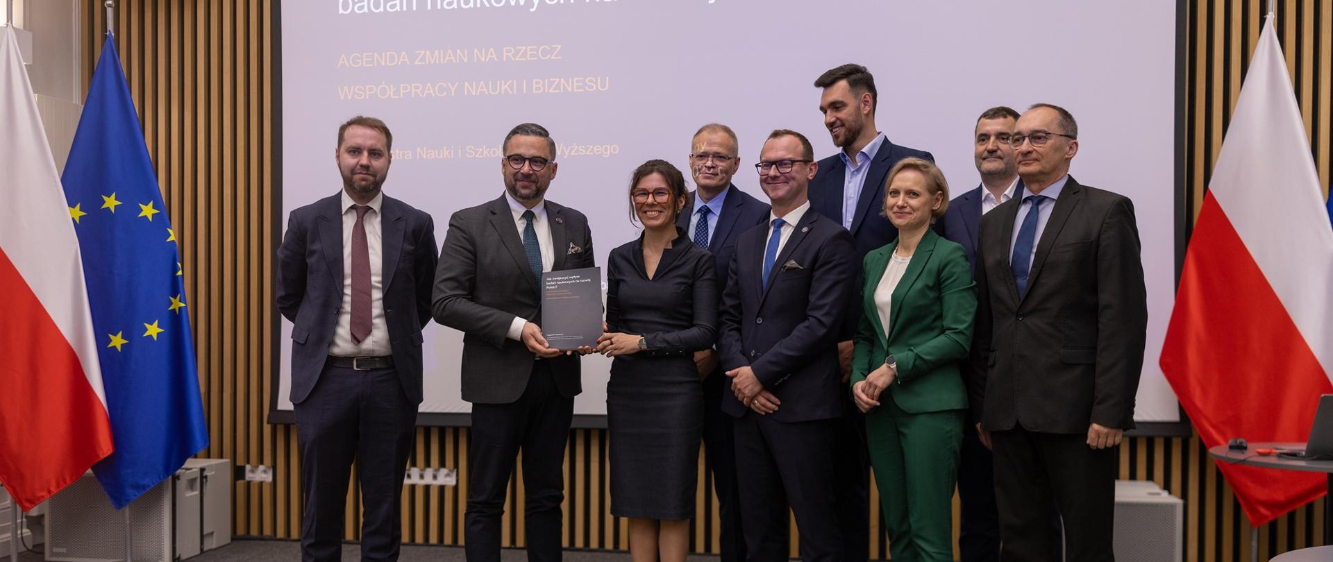 Pod wbudowanym w ścianę z cienkich pionowych listewek wielkim ekranem stoi dziewięć osób, drugi od lewej minister Kulasek trzyma szarą teczkę, za nimi pod ścianą po obu stronach flagi Polski i UE.