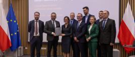 Pod wbudowanym w ścianę z cienkich pionowych listewek wielkim ekranem stoi dziewięć osób, drugi od lewej minister Kulasek trzyma szarą teczkę, za nimi pod ścianą po obu stronach flagi Polski i UE.