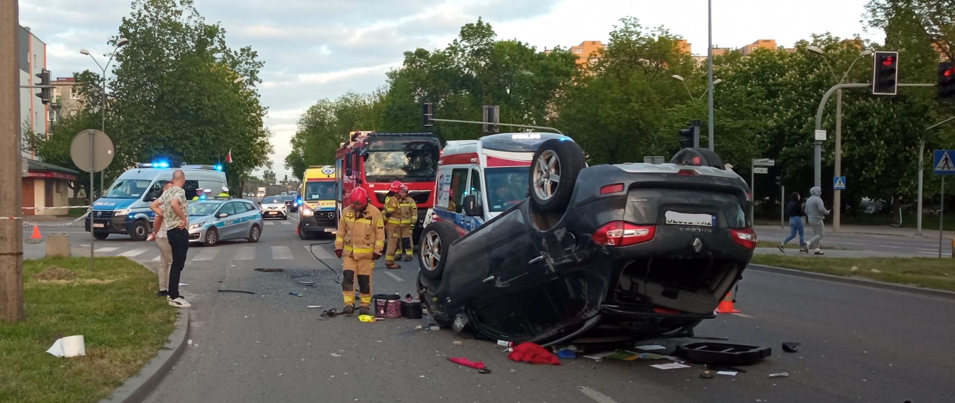 Zdjęcie prezentuje szeroki kadr z miejsca wypadku samochodowego. W centralnym punkcie samochód Renault przewrócony na dach. Za autem stoją samochody służb ratowniczych: 2 radiowozy, 2 karetki i pojazd pożarniczy. Przy uszkodzonym aucie stoją strażacy.