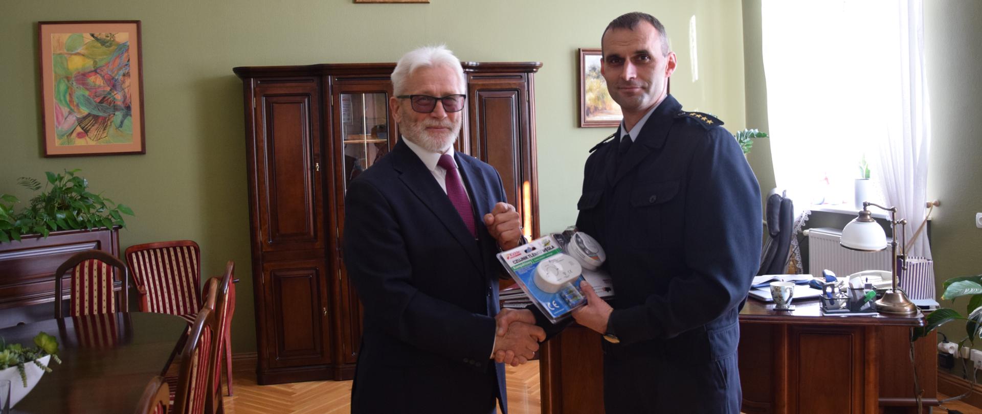 KOLEJNY ROK POWIAT FUNDUJE CZUJKI DLA MIESZKAŃCÓW. Na zdjęciu stoją dwie osoby - Starosta Kościański i Komendant Powiatowy PSP - symbolicznie przekazywane są czujki tlenku węgla i czujki dymu, w tle biuro.