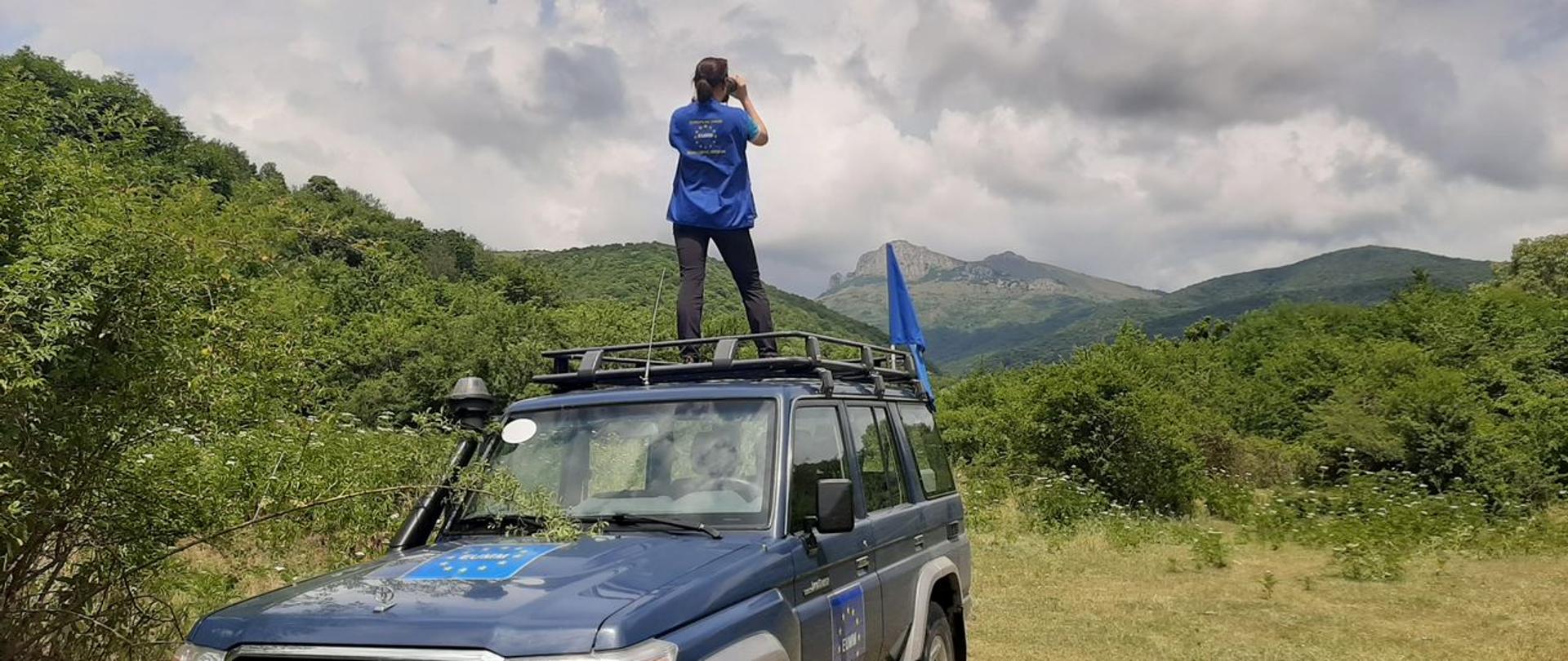 Kobieta na dachu samochodu, patrzy przez lornetkę.