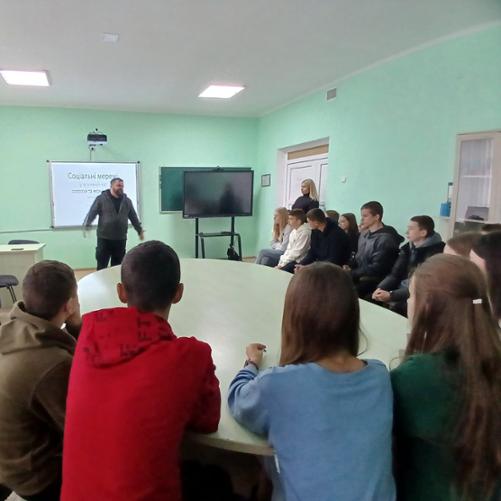 W sali grupa nastolatków siedzi wokół owalnego stołu i słucha prowadzącego szkolenie, który stoi obok tablicy, ekranu projektora i telewizora na stojaku