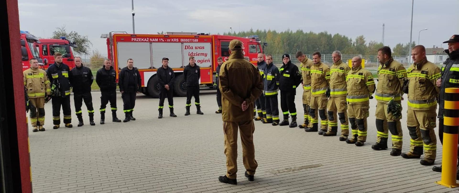 Zdjęcie przedstawia strażaków podczas zbiórki stojących w kwadracie, za nimi stoją wozy gaśnicze, w środku odwrócony tyłem Komendant