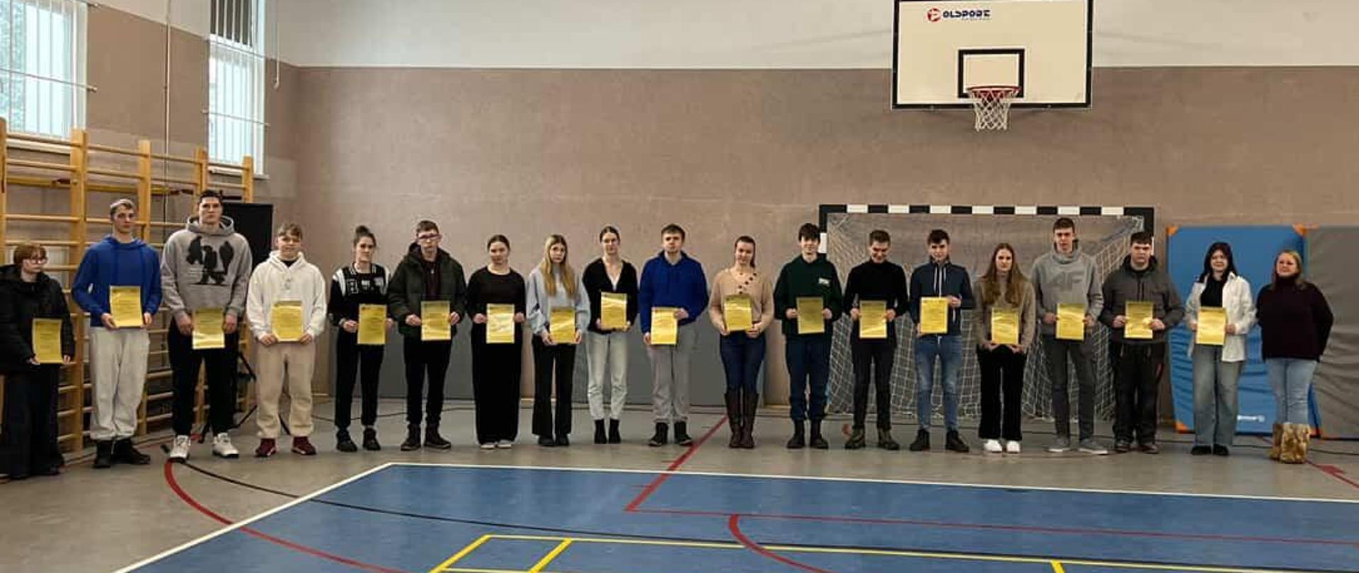 Liczna grupa młodzieży stoi w jednym rzędzie na boisku do koszykówki, prezentując otrzymane dyplomy.