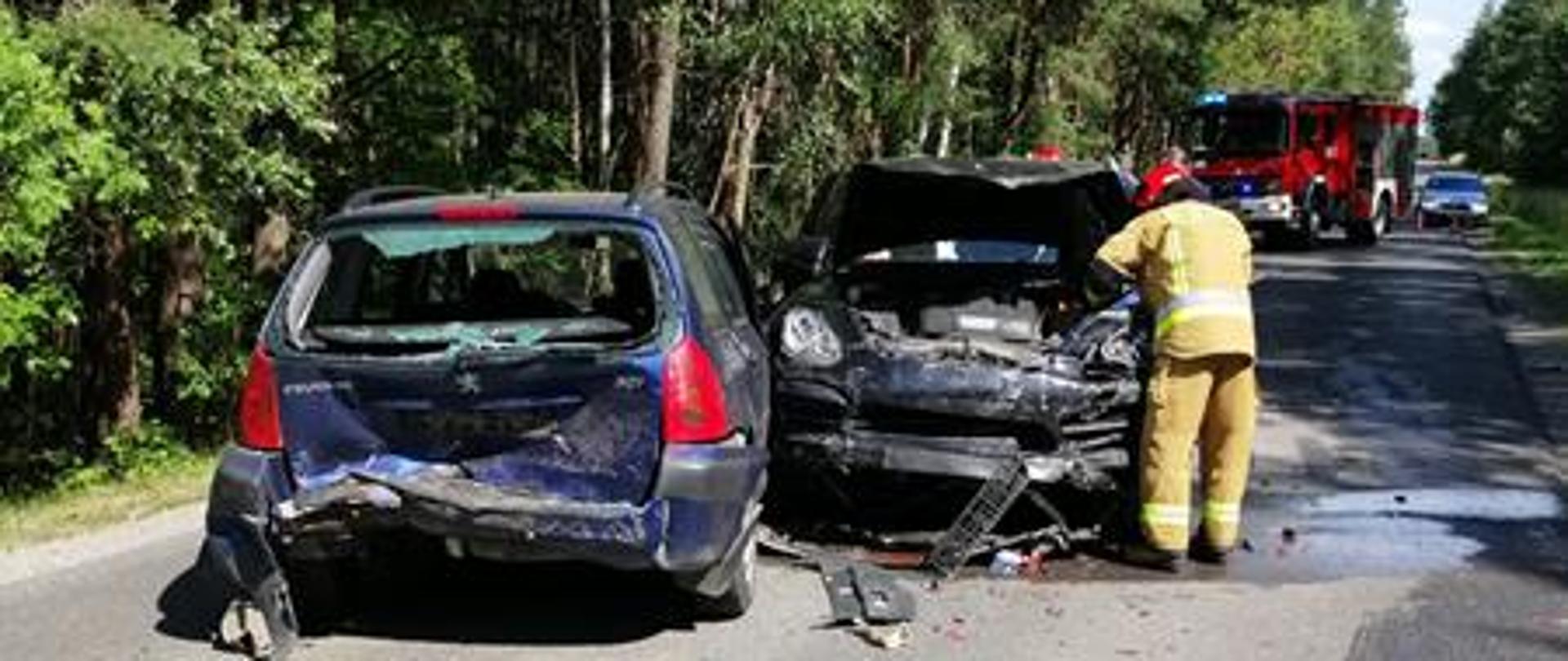 Na drodze znajdują się dwa pojazdy, przy których strażak ubrany w ubranie specjalne prowadzi działania. Na drugim planie widać samochód pożarniczy z włączonymi sygnałami świetlnymi.