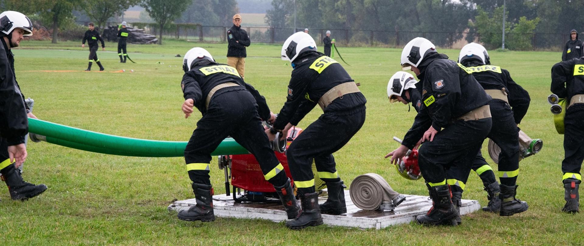 Drużyna męska OSP podczas dobiegnięcia do motopompy, zabierająca sprzęt z podestu.