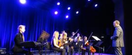 Zdjęcie przedstawia trwający koncert i muzyków grających na skrzypcach oraz keybordzie, nie brakuje też dyrygenta. 