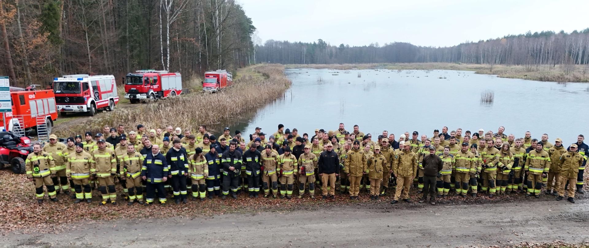 Duża grupa strażaków ustawiona jest w szerokim półkolu na leśnej drodze. Wewnętrzną część kręgu zajmują prowadzący i przedstawiciele służb. Otoczenie stanowi jesienny las, z drzewami o bezlistnych koronach i warstwą opadłych liści na ziemi. Z góry widać skalę wydarzenia oraz liczbę uczestników biorących udział w ćwiczeniach „Kozieławy 2025”.