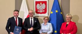 4 elegancko ubrane osoby, pozują do zdjęcia, na tle flag UE i Polski oraz orła