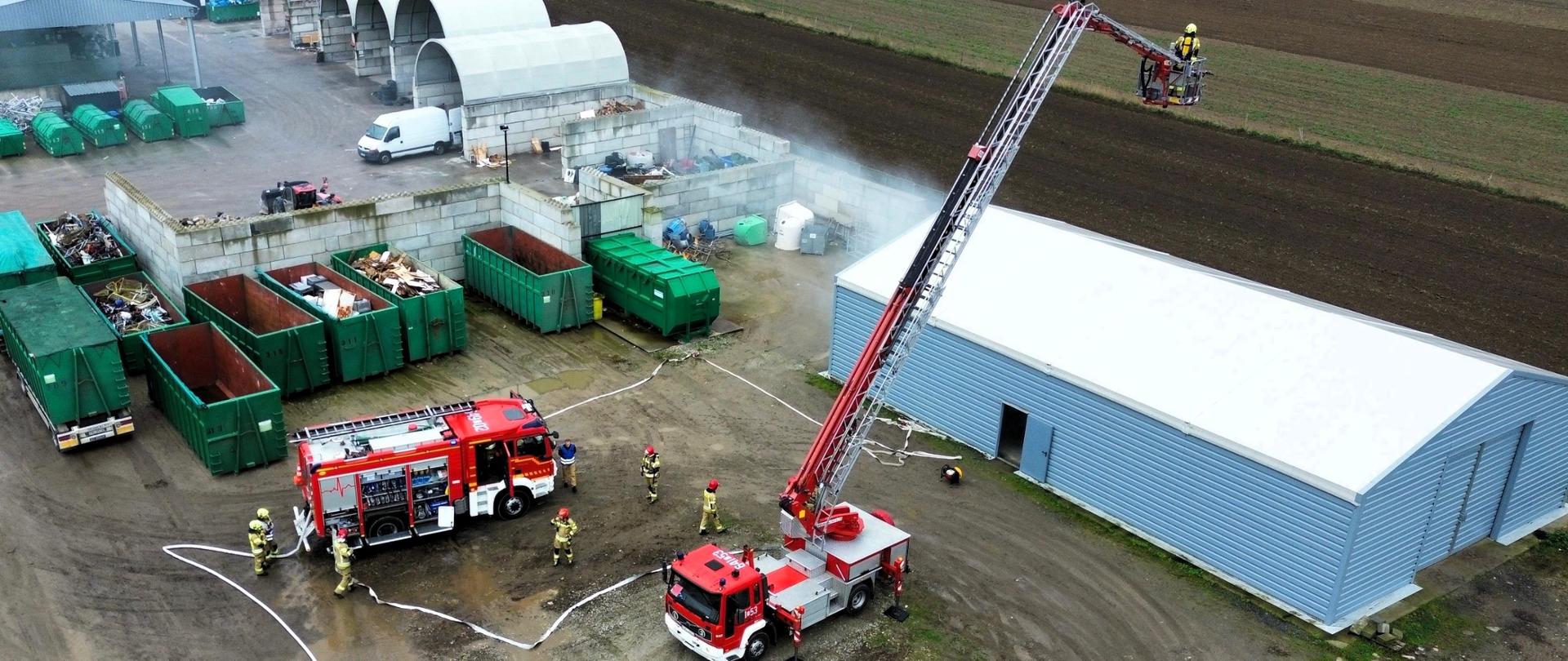 Na placu manewrowym stoją dwa wozy strażackie – jeden z drabiną mechaniczną, drugi to wóz gaśniczy z rozwiniętymi wężami. Kilku strażaków w ubraniach bojowych pracuje przy pojazdach – jedni podłączają węże, inni sprawdzają sprzęt. Na ziemi widać błoto i wodę, a niebo jest zachmurzone, co sugeruje, że ćwiczenia odbywają się w deszczową pogodę.