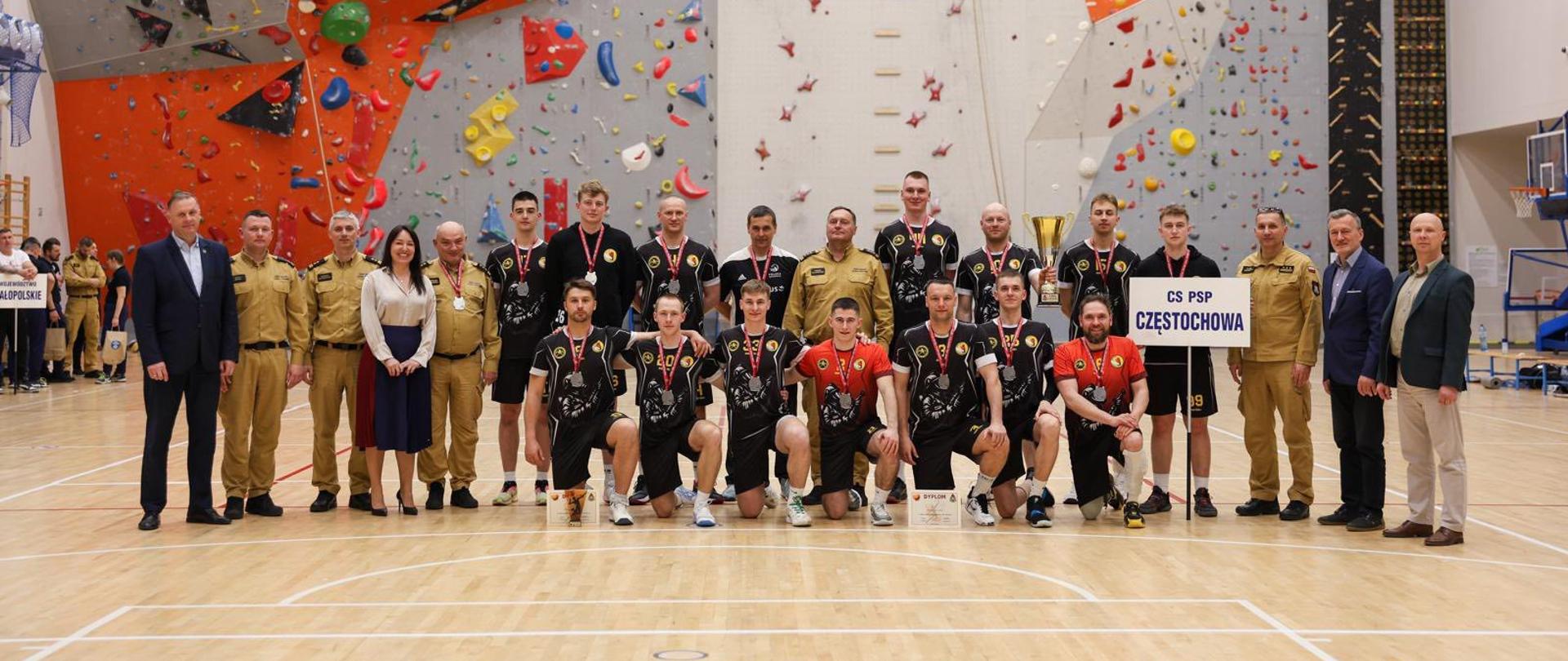 Pamiątkowe zdjęcie drużyny Centralnej Szkoły PSP. Zawodnicy z pucharem i medalami po zakończeniu turnieju w towarzystwie osób zaangażowanych w organizację mistrzostw oraz przedstawicieli PSP