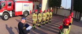 Na pierwszym planie strażacy w dwóch rzędach stoją podczas zmiany służbowej. W tle wóz strażacki przed budynkiem straży.