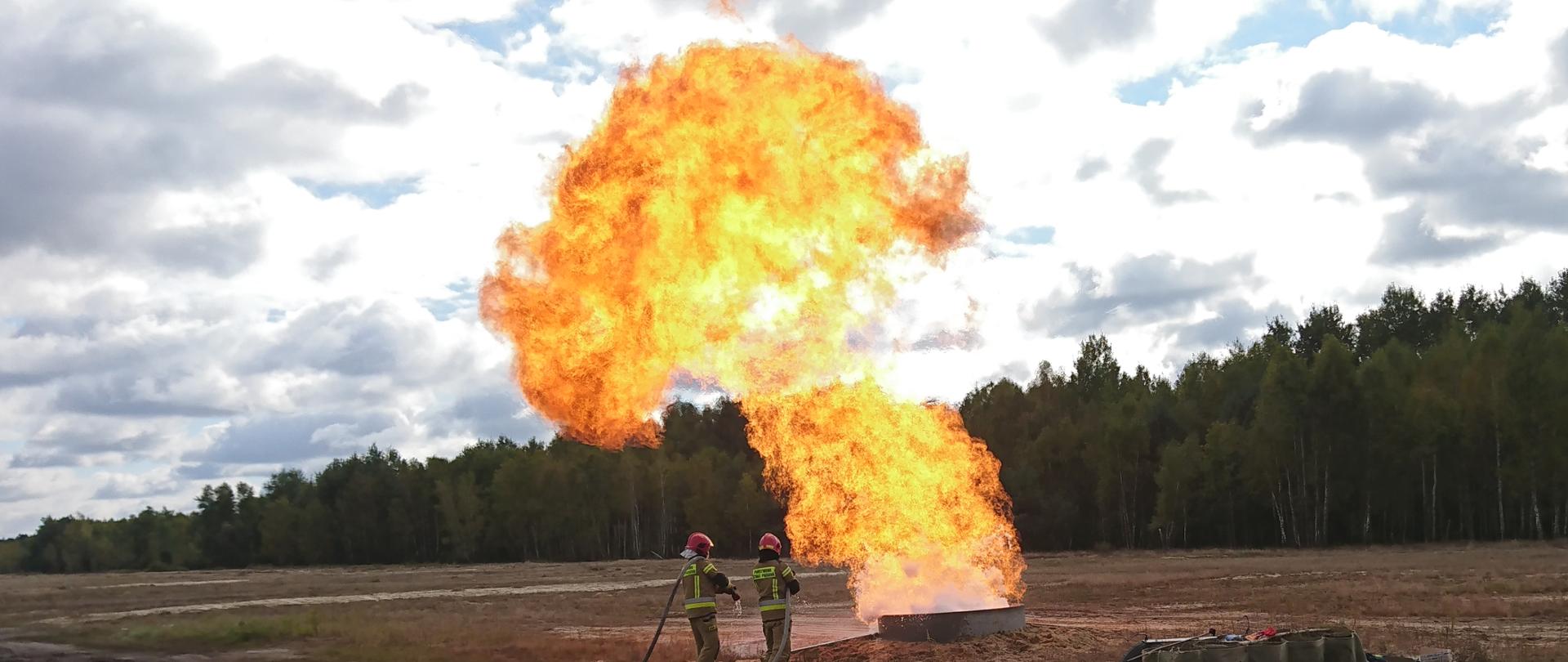 Zdjęcie przedstawia gaszenie pożaru rozlewiska gazu LNG. Miejsce zdarzenia to gruntowa przestrzeń otwarta. W oddali po prawej stronie znajduje się las. Rozlewisko gazu znajduje się betonowej studni wystającej na powierzchnie ziemi około pól metra oraz średnicy około 2 metrów. Ogień unosi się na wysokość około dziesięciu metrów. W gaszeniu pożaru bieżę udział dwóch strażaków oddaleni od źródła ognia około trzech metrów ubrani w ubrania specjalne podając dwa prądy wody w natarciu na źródło ognia. W dolnej części zdjęcia po prawej stronie oddalony około 20 metrów od źródła ognia znajduje się Oficer obserwujący działania gaśnicze. 