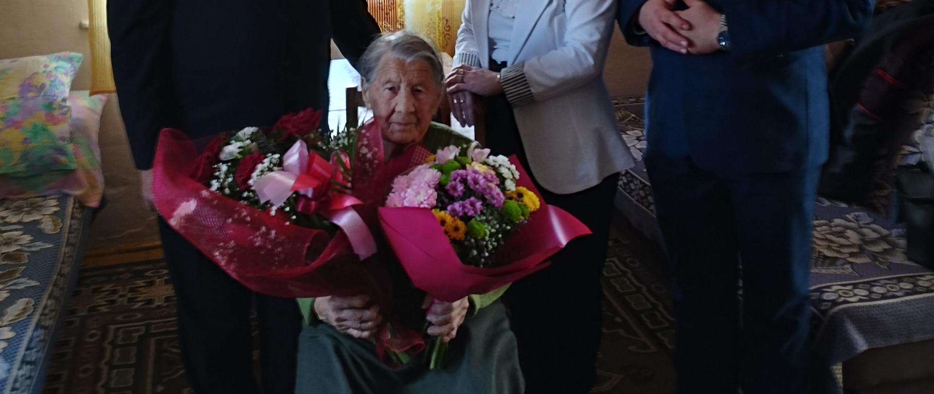 Na krześle siedzi starsza kobieta z dwoma ogromnymi bukietami kwiatów. Jest bardzo przejęta życzeniami i gratulacjami. Za nią po lewej stronie stoi lekko nachylony w kierunku starszej pani mężczyzna, a po prawej stronie stoją kobieta w białej marynarce i mężczyzna w garniturze.