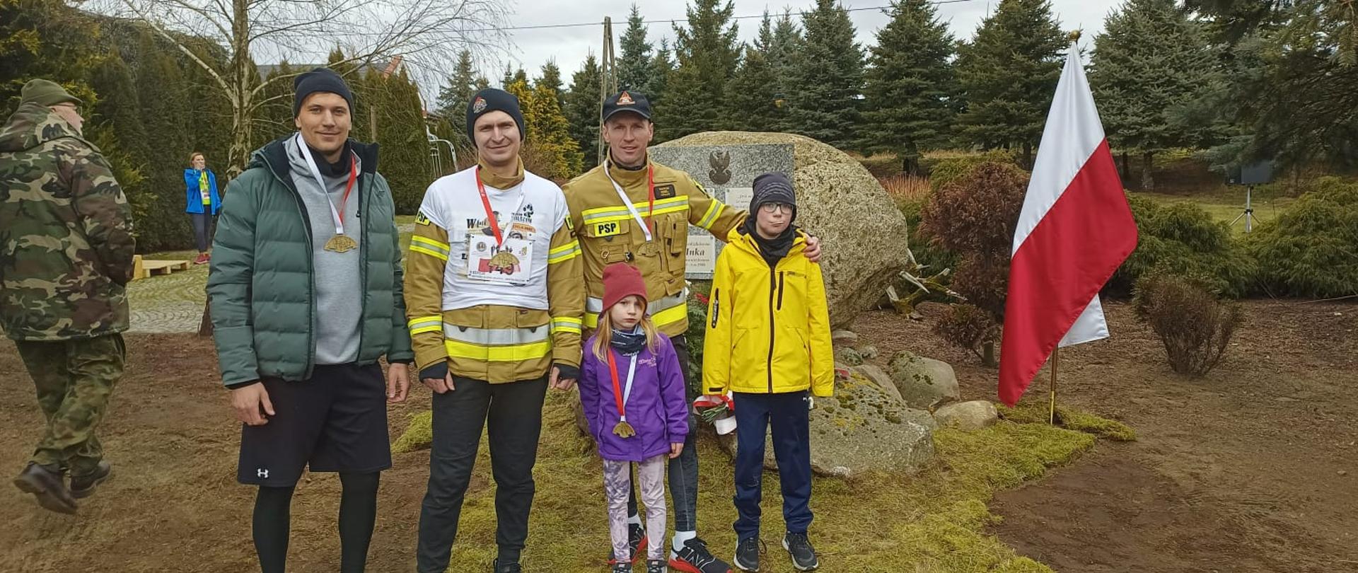 Zdjęcie przedstawia uczestników biegu z medalami na szyi stojącymi za kamieniem oraz flaga Polski.