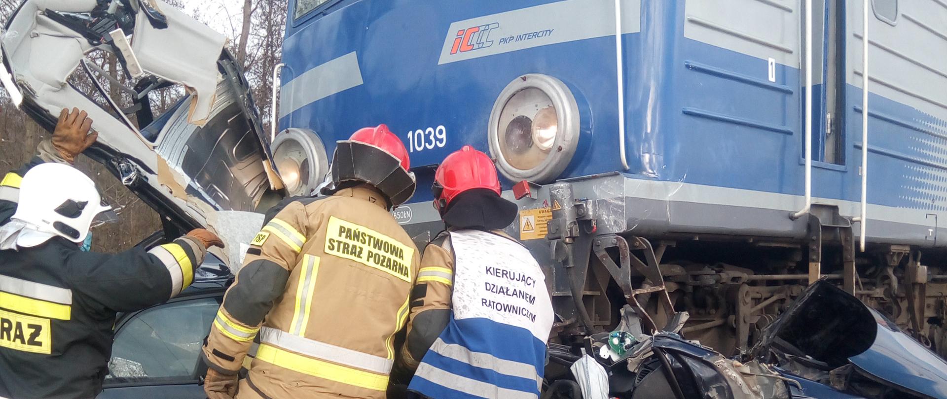 Na zdjęciu widać trzech strażaków pracujących przy akcji ratowniczej. Niebieski pociąg zmiażdżył samochód a jego elementy wystają z pod pociągu.