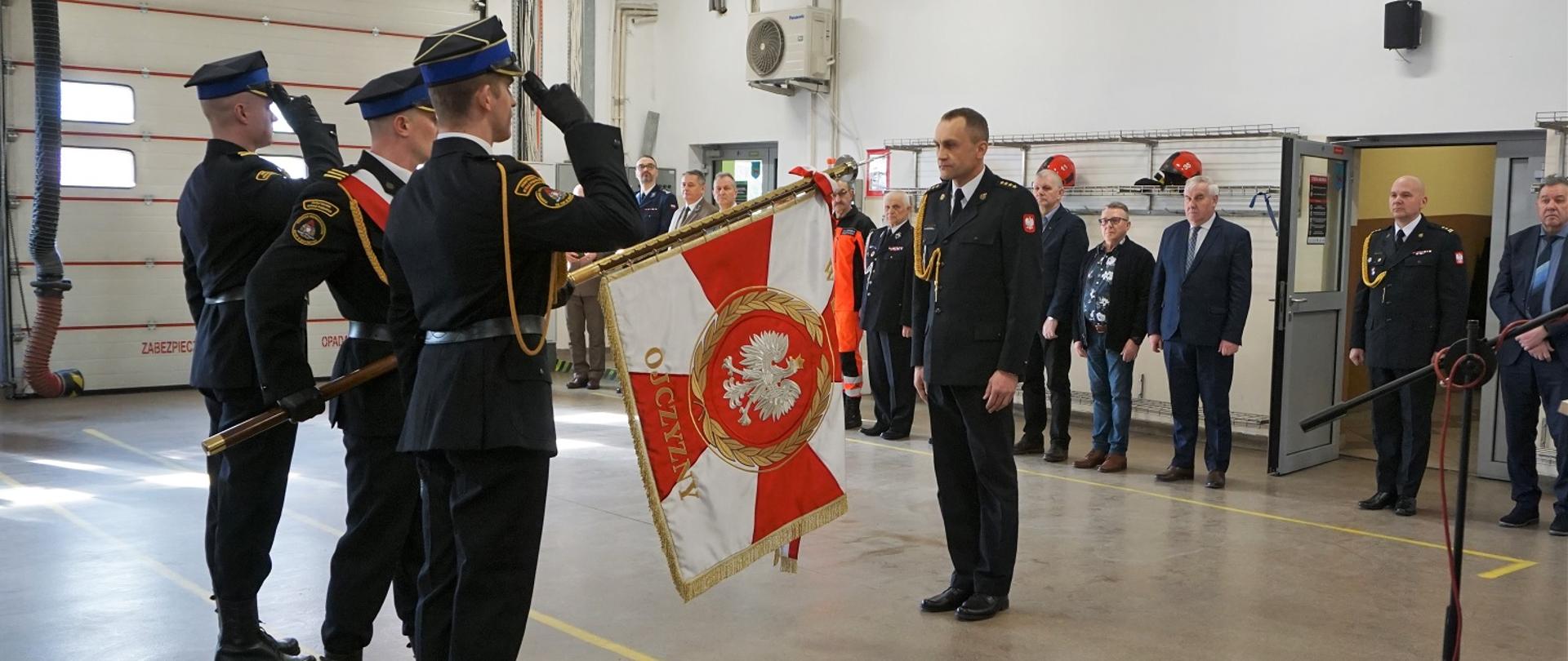 Nowy Komendant Powiatowy PSP w Grajewie stoi przed sztandarem komendy. Poczet salutuje. W tle zaproszeni goście. 